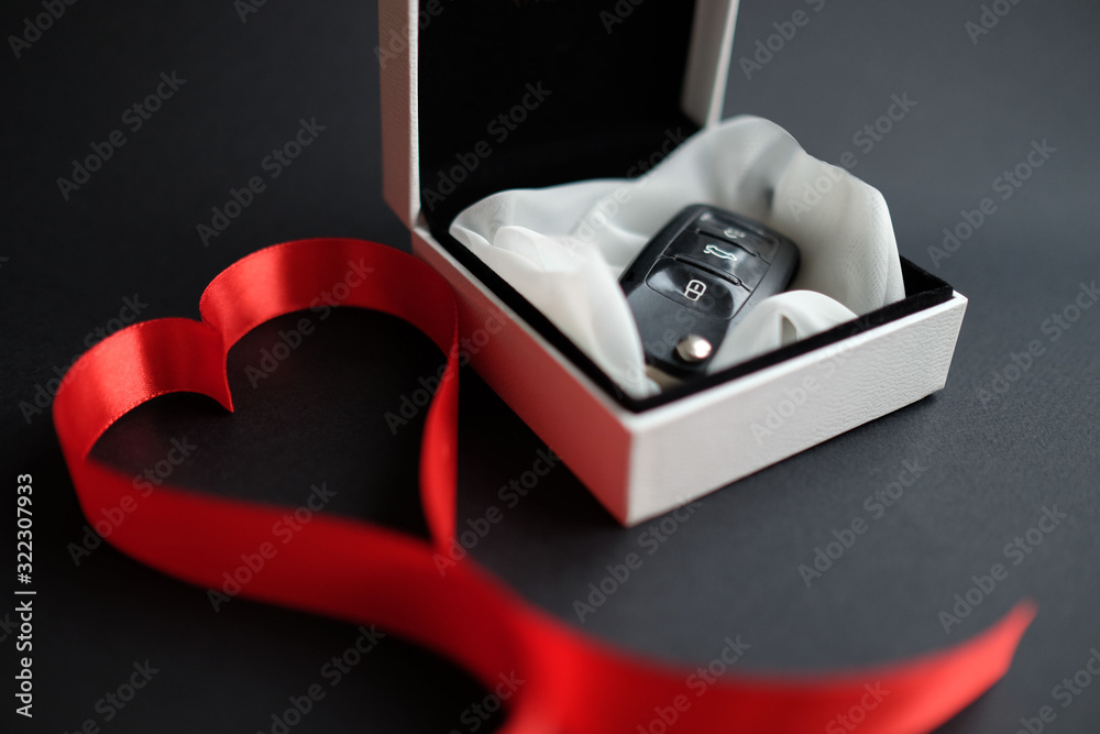 White gift box with car keys with remote control alarm system with red ribbon in the shape of a heart. St Valentine holiday concept. White gift box with car keys with remote control alarm system with red ribbon in the shape of a heart. St Valentine holiday concept.