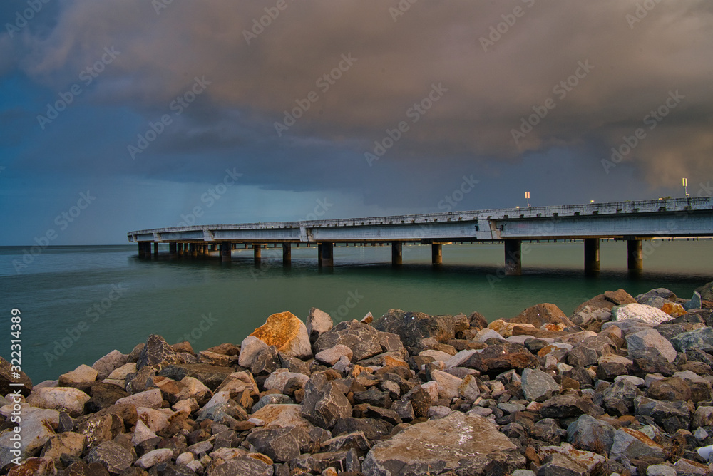 Eine menschenleere Brücke über das Meer in Panama, Steine im Vordergrund und lila Wolken, kurz vor einem Gewitter