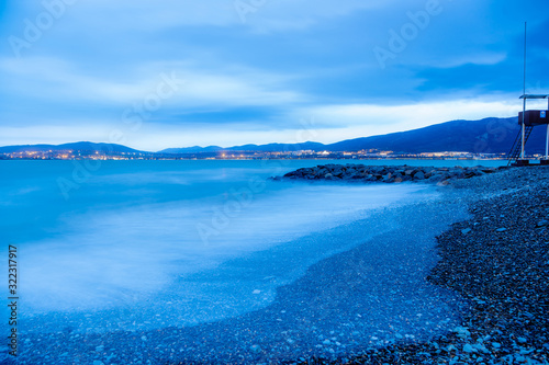 Gelendzhik beach in the evening in a storm. The waves at high speed turn into a blue fog. Blue tones, winter. Pebble beach, breakwater. In the background, the Caucasus mountains. 