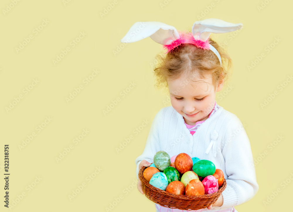 Cute blonde 5 year old girl with funny bunny ears costume, holding wicker basket with colorful Easter eggs on yellow background.