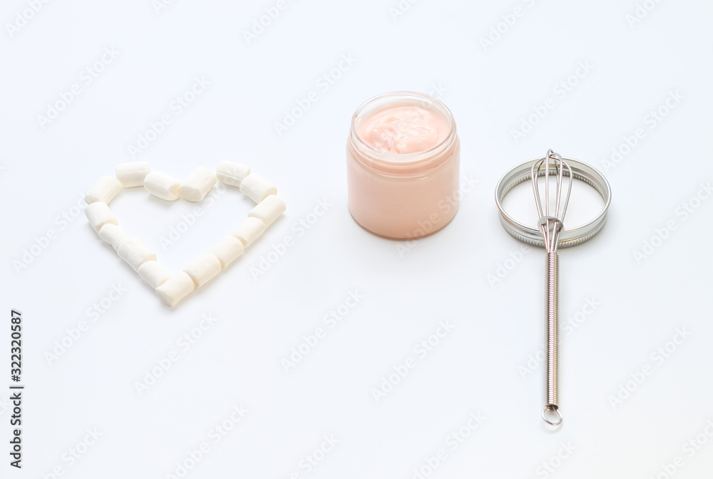 a glass transparent jar with a pastel peachy cream in an opened jar with a metal top and a clean metal whisk lying with a heart, concept of enjoying using and making handmade cosmetics