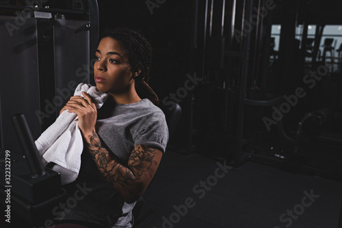 tattooed african american woman holding towel near fitness machine