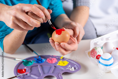 Mother and her child hands painting Easter eggs. The mother helps her son paint an egg with a paintbrush at home. Happy family preparing for Easter. Easter holiday concept. Close up, selective focus