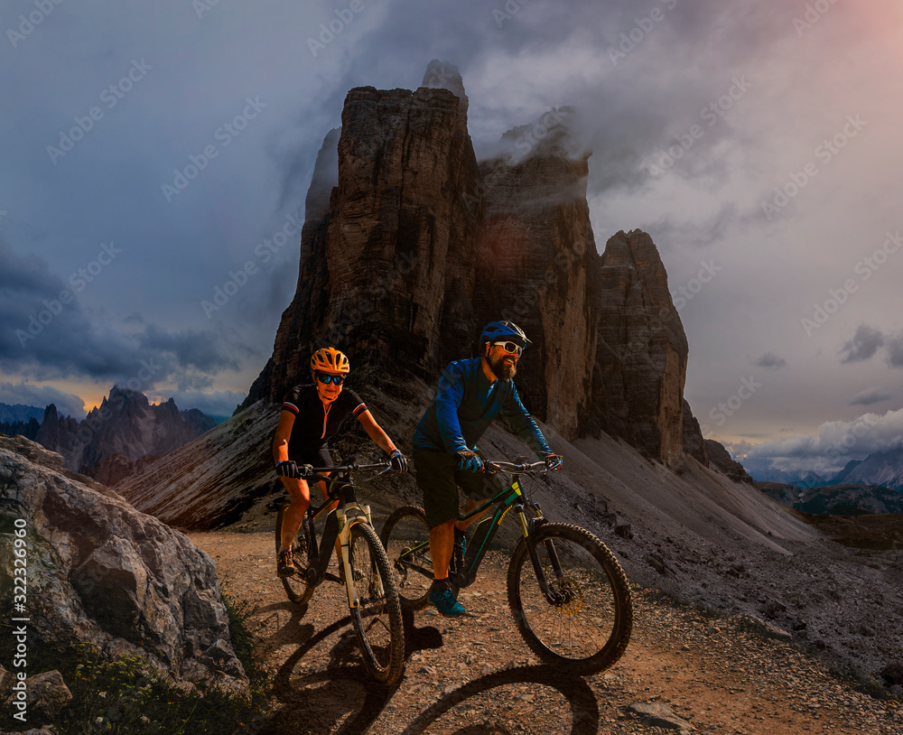 Fototapeta premium Couple cycling on electric bike, rides mountain trail. Woman and Man riding on bikes in Dolomites mountains landscape. Cycling e-mtb enduro trail track. Outdoor sport activity.