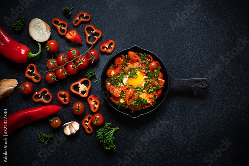 Shakshouka eggs poached in a sauce of tomatoes, vegetables, spices and herbs in iron pan. Bread, tomatoes, red peppers, garlic, parsley. Traditional breakfast. Black background, top view, copy space