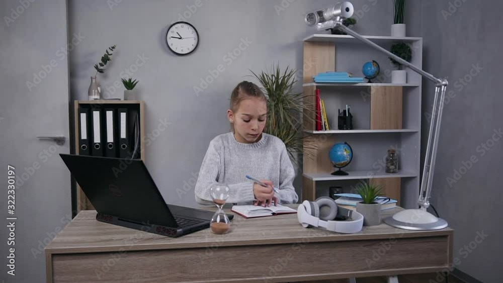 Lovely concentrated teen girl with long light hair sitting at her home workplace and working with computer and notebook Lovely concentrated teen girl with long light hair sitting at her home workplace and working with computer and notebook