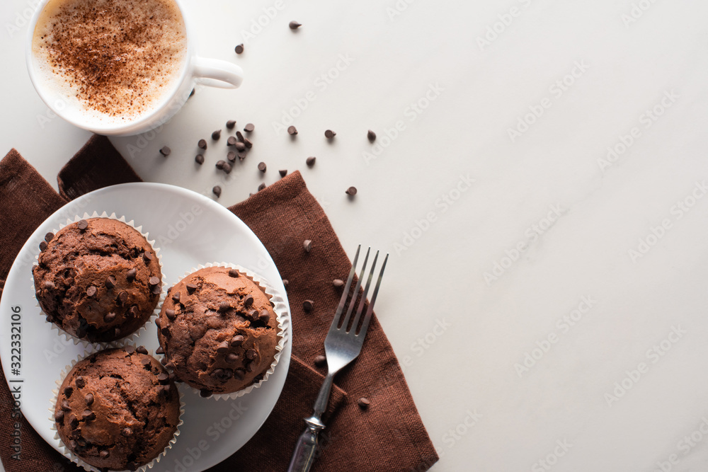 top view of fresh chocolate muffins on white plate and brown napkin near fork and cappuccino on marble surface top view of fresh chocolate muffins on white plate and brown napkin near fork and cappuccino on marble surface
