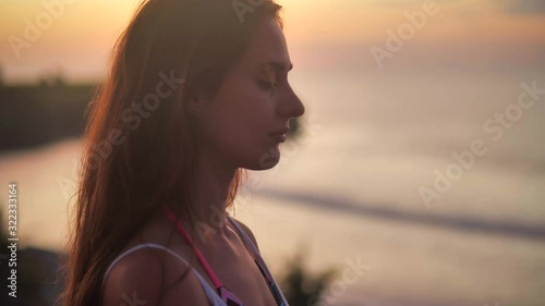 Beautiful girl praying looking up at orange sky with hope, close-up. Silhouette of young woman dreaming looking upwards sunset outdoors ocean background 4k.