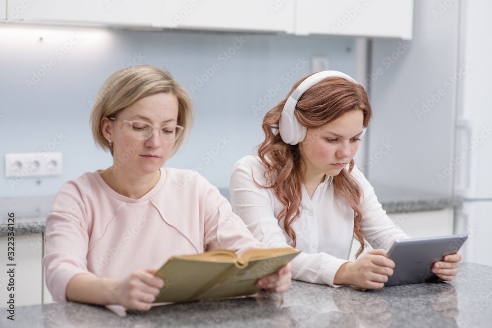 A teenage girl sitting with a tablet computer in her hands and in headphones, next to her mother is sitting in glasses and reading a book. In the kitchen in your home A teenage girl sitting with a tablet computer in her hands and in headphones, next to her mother is sitting in glasses and reading a book. In the kitchen in your home