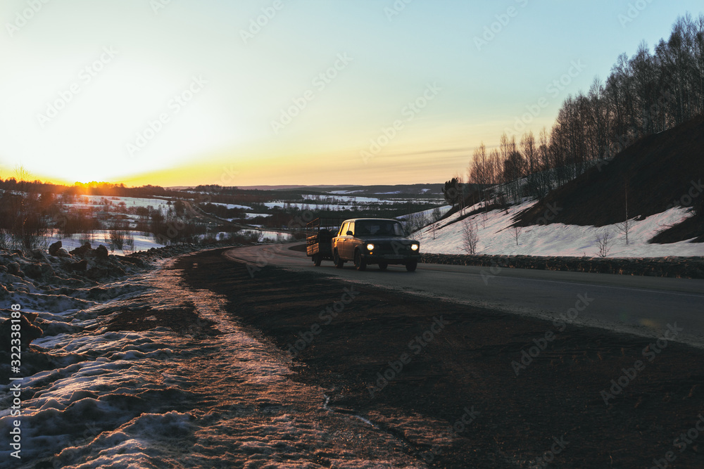 A car with a trailer rides on a winter road in the rays of the setting sun