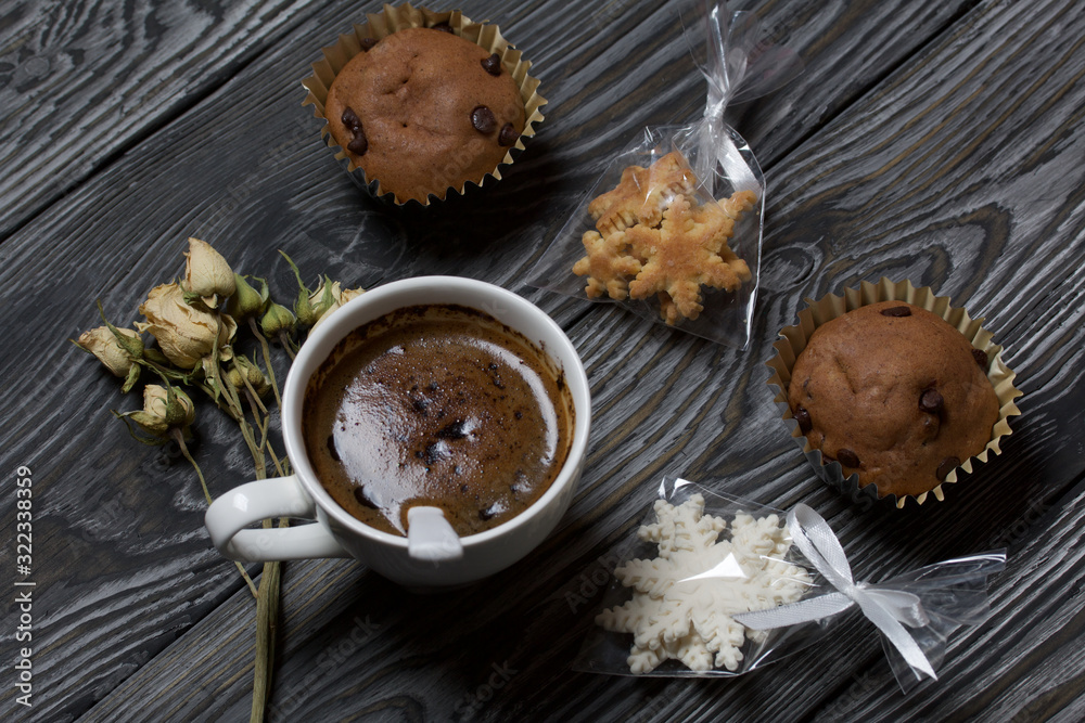 Coffee in a white cup. Nearby are muffins, and a dried rose of beige color. Sachets of cookies in the shape of snowflakes are visible. On brushed pine boards. Coffee in a white cup. Nearby are muffins, and a dried rose of beige color. Sachets of cookies in the shape of snowflakes are visible. On brushed pine boards.