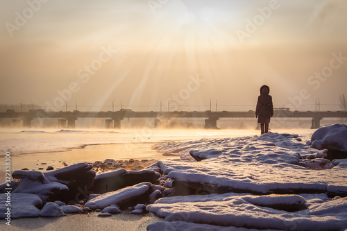 A woman meets the winter dawn near the river near the bridge
