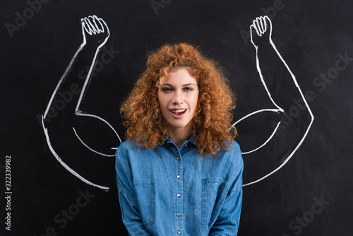 portrait of smiling redhead woman with strong muscular arms drawing on blackboard