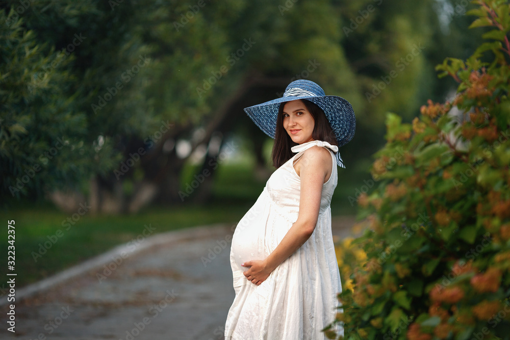 Young beautiful pregnant girl of Caucasian appearance in white dress and hat in nature in the Park.