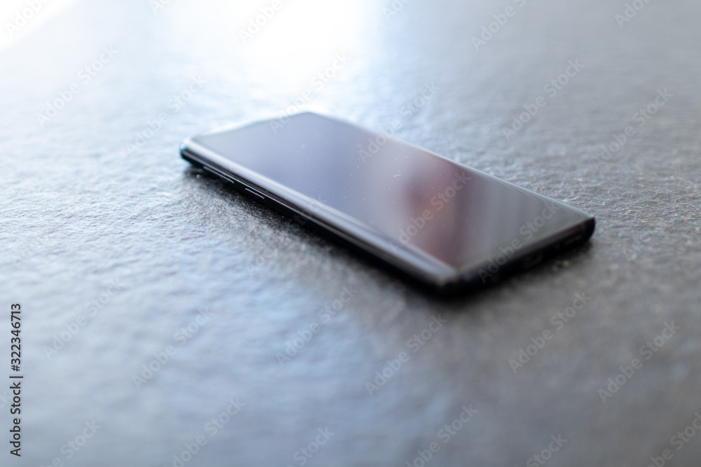 Smartphone on table. Glossy black smartphone on an illuminated surface. Digital communication business tools. Smartphone and social media tools with glare bright screen on natural light environment. Smartphone on table. Glossy black smartphone on an illuminated surface. Digital communication business tools. Smartphone and social media tools with glare bright screen on natural light environment.