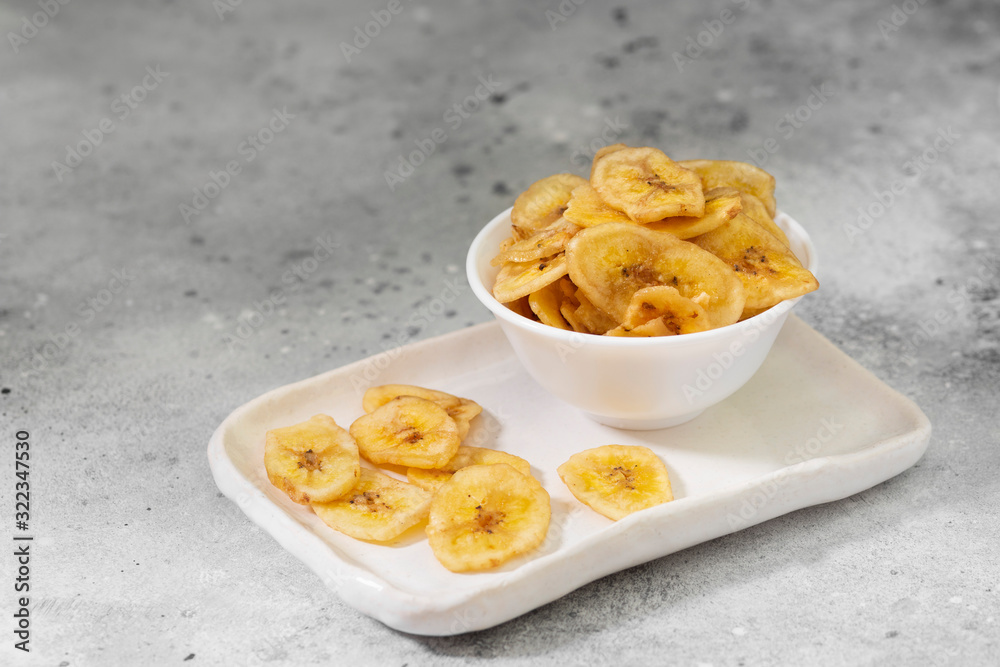 Dried bananas in a white ceramic bowl on a gray kitchen table. Banana chips. Vegetarian snack for proper nutrition Dried bananas in a white ceramic bowl on a gray kitchen table. Banana chips. Vegetarian snack for proper nutrition