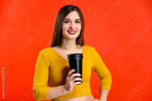 nice brunette girl with long hair with a smile with a cup of coffee in her hands in a yellow sweater is happy over a red background smiling and showing positive emotions