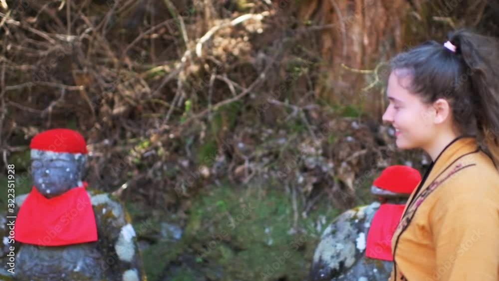 Slow motion side shot of young woman profile walking, looking at row of Jizo statues in Kanmangafuchi Abyss in Tochigi prefecture in Nikko, Japan in early spring with moss