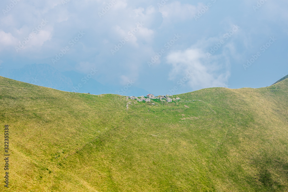 Old Stone abandoned houses on a green alpine ridge. Old ruined farm house in the mountains above Lake Como in Italy, blue sky with clouds in the background. Old Stone abandoned houses on a green alpine ridge. Old ruined farm house in the mountains above Lake Como in Italy, blue sky with clouds in the background.