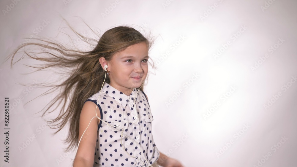 Child with earflaps dancing at studio background. The girl listens to music on the smartphone. The kid has loose long hair. Beautiful cute happy little girl listens to music on Headphones. Child with earflaps dancing at studio background. The girl listens to music on the smartphone. The kid has loose long hair. Beautiful cute happy little girl listens to music on Headphones.