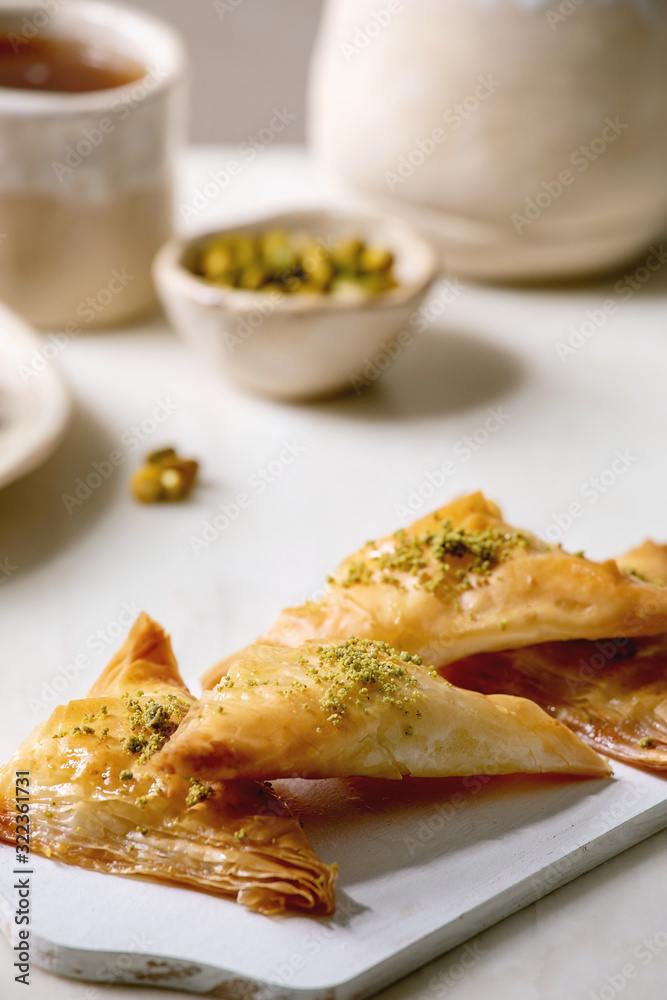 Homemade Turkish traditional dessert baklava with pistachio served on white wooden cutting board with bowl of nuts, cup of tea, ceramic teapot over white marble table. Homemade Turkish traditional dessert baklava with pistachio served on white wooden cutting board with bowl of nuts, cup of tea, ceramic teapot over white marble table.