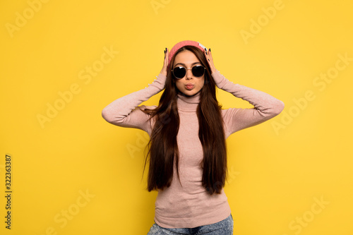 Indoor studio shot of trendy stylish woman with long dark hair wearing pink cap and black glasses over yellow background. Front view of happy girl isolated on yellow background.