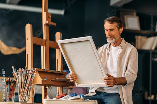 Senior painter sitting in his creative studio and holding a canvas with a picture