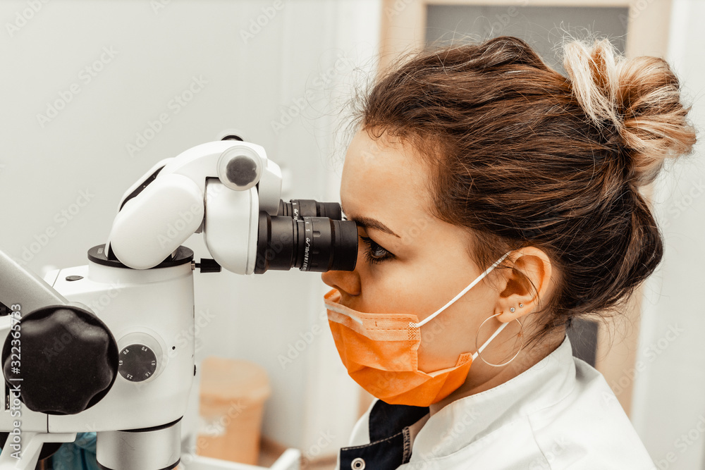 Young woman dentist doctor looks through a professional microscope in a dental clinic. A doctor in a disposable medical mask and cap. Advanced equipment in dentistry Young woman dentist doctor looks through a professional microscope in a dental clinic. A doctor in a disposable medical mask and cap. Advanced equipment in dentistry