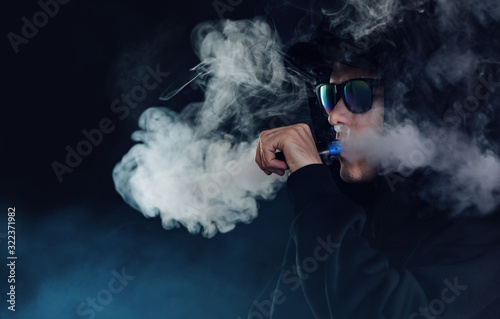 vape man. portrait of a handsome guy in a black cap and glasses vaping and exhaling a cloud of vapor from an e cigarette dark background