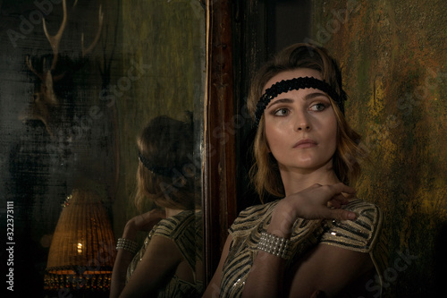 Portrait of young adult beautiful female model in 1920s style golden pattern dress looking away from huge mirror. Roaring twenties gatsby style model concept. Dramatic lighting indoor photoshoot.