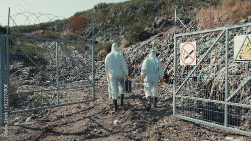 Ecologists in protective clothes walking and observing huge landfill site. People scientists working on trash samples in toxic area. photo