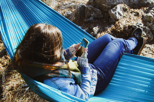 The girl is resting in a turquoise hammock and uses the phone on vacation.