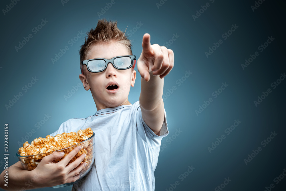 The boy holds popcorn in his hands watching a movie in 3D glasses on a blue background. The concept of a cinema, films, emotions, surprise, leisure. The boy holds popcorn in his hands watching a movie in 3D glasses on a blue background. The concept of a cinema, films, emotions, surprise, leisure.