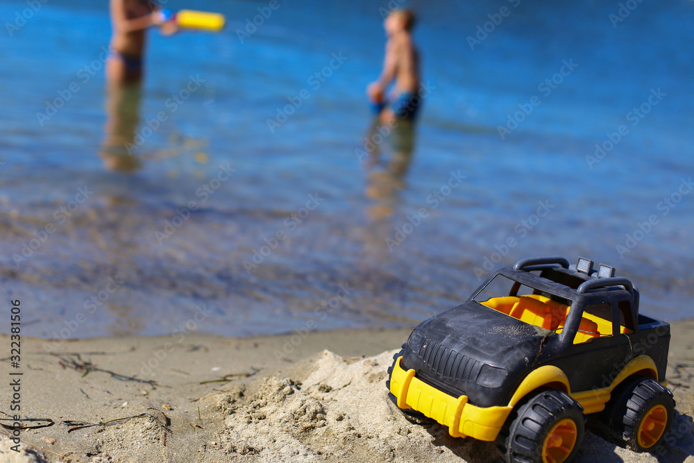 Obraz premium Children toy car jeep of black and yellow color on sand near the water on the beach of blue sea. Boys play water guns. Vacation with kids on coast on warm sunny summer day. Tourism in Europe,Slovenia