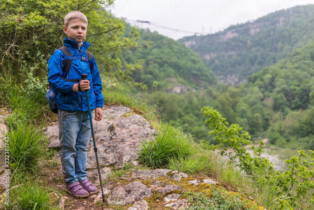 A boy with a backpack and trekking poles stands on a stone rock at the beginning of a mountain gorge