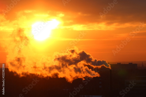 Red sun at sunset or dawn at dusk low above the horizon shrouded in smoke from the chimneys of the plant