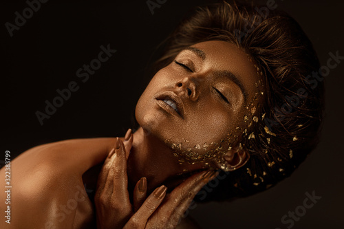 Fashion art portrait of model girl with holiday golden shiny professional makeup. beaty woman with gold metallic body and hair on dark background. Gold glowing skin. photo