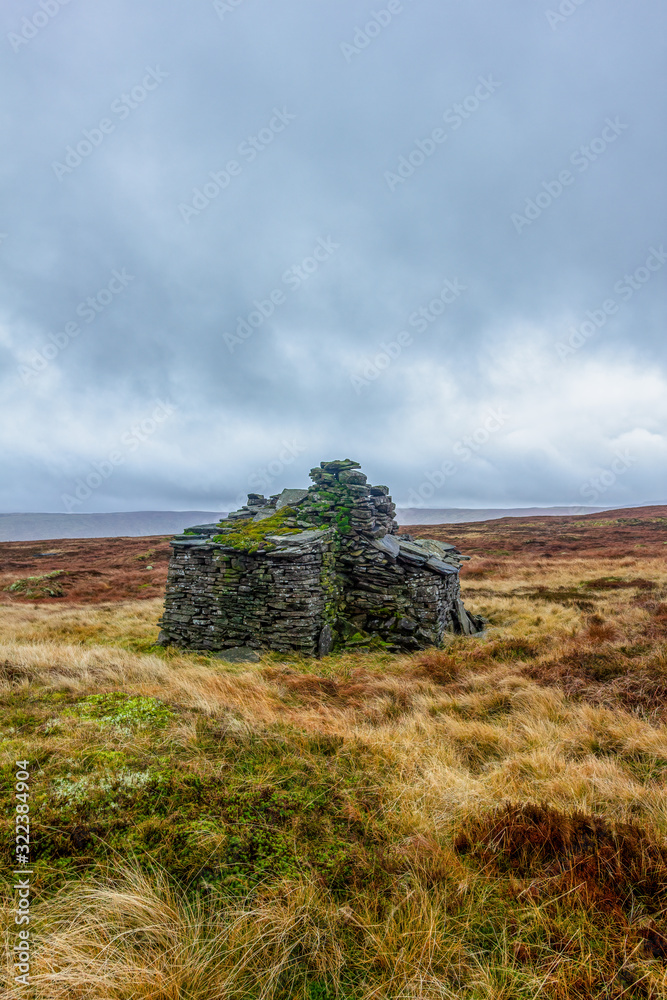 A scenic view of a stony mountain shelter in ruin surrounded with grass and bog under a grey sky