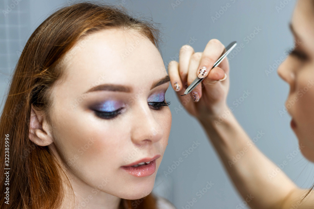 Obraz premium Skillful visagiste in white t-shirt is holding brush, applying make-up for young model while posing sideways against gray studio background. Close up