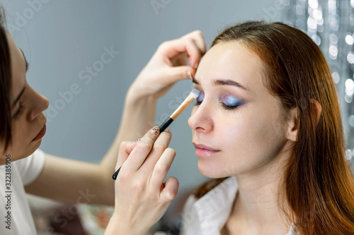 Skillful visagiste in white t-shirt is holding brush, applying make-up for young model while posing sideways against gray studio background. Close up