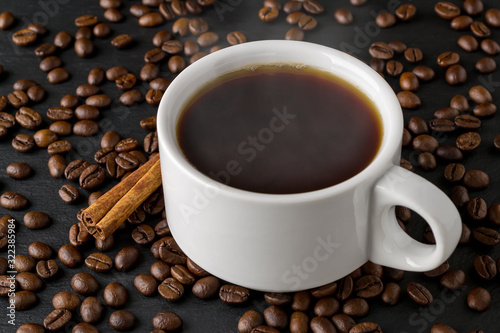 Hot coffee in white elegant cup and cinnamon stick on a background of roasted coffee beans over a black stone serving board. Coffee with cinnamon and anise.