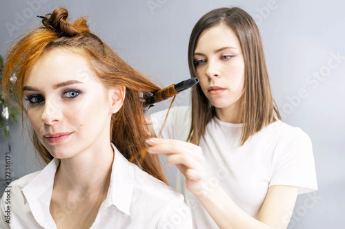 Hairdresser in white t-shirt is spraying hair of young redhead model with bright make-up with liquid from a bottle, making a hairstyle. Close up