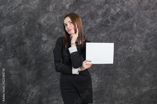 Brunette woman in black dress, hand on hip, holding white empty blank board ready for your text or product, posing on gray background. Close up