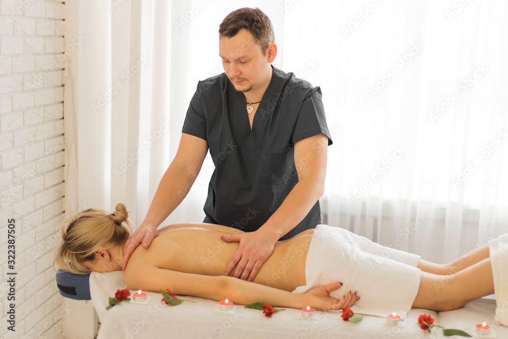 Hands of man masseur making therapeutic back massage for girl lying on couch in spa salon. Flowers and burning candles are nearby. Close up Hands of man masseur making therapeutic back massage for girl lying on couch in spa salon. Flowers and burning candles are nearby. Close up