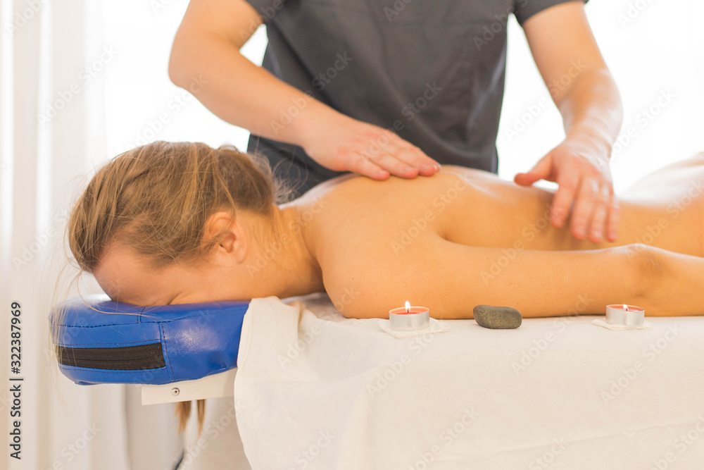 Hands of masseur making therapeutic back massage for female lying on couch in private clinic. Flowers and burning candles are nearby. Close up Hands of masseur making therapeutic back massage for female lying on couch in private clinic. Flowers and burning candles are nearby. Close up