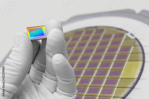 Hand in white glove holding microchip photo sensor matrix. On the background is diced silicon wafer with microchips. Focus on chip.
