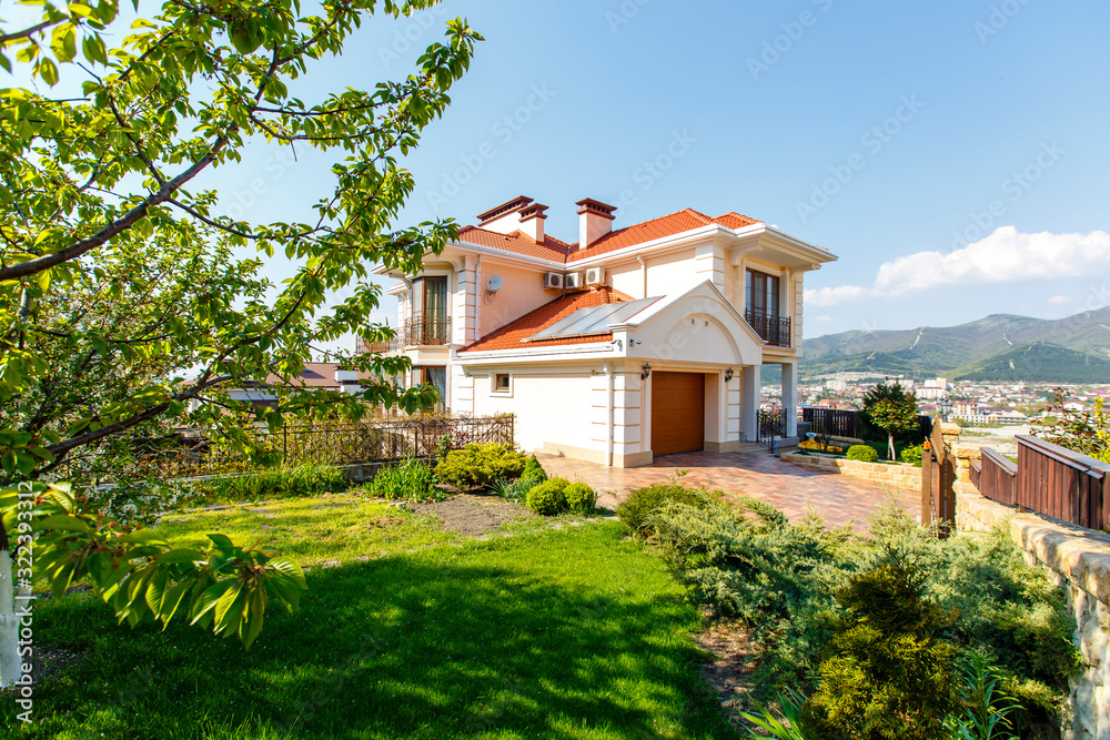 The facade of a classic Mediterranean two-storey cottage in the spring. In front of the cottage there is a green lawn and flowering trees. Mountains in the background. The facade of a classic Mediterranean two-storey cottage in the spring. In front of the cottage there is a green lawn and flowering trees. Mountains in the background.