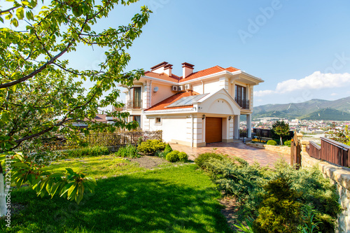 The facade of a classic Mediterranean two-storey cottage in the spring. In front of the cottage there is a green lawn and flowering trees. Mountains in the background.