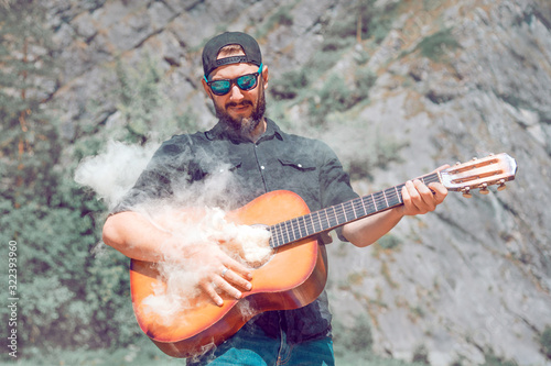 a man in sunglasses in a black shirt plays the guitar from which smoke comes, against the background of nature