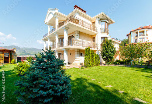 The backyard of a classic three-story white cottage with wrought-iron railings and columns. In front of the cottage there is a green lawn, trees and an empty pool.  © Александр Трихонюк